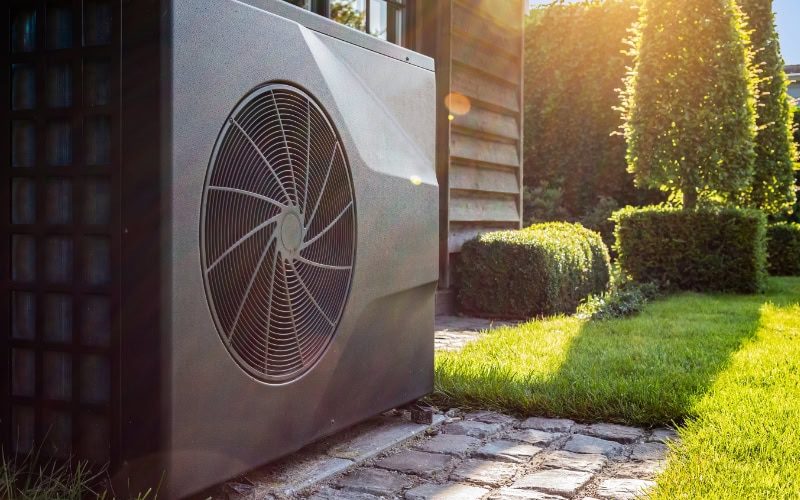 Transitioning Your Heat Pump. Close-up of black full inverter heat pump outside in the garden, near wooden pool house on a sunny day. Lens flare on the image.