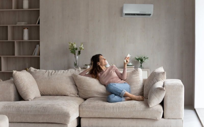 Planning to Remodel? Go Ductless! Relaxed young hispanic female homeowner sitting on huge comfortable couch, turning on air conditioner with remote controller, switching on cooler system, setting comfortable temperature in living room.