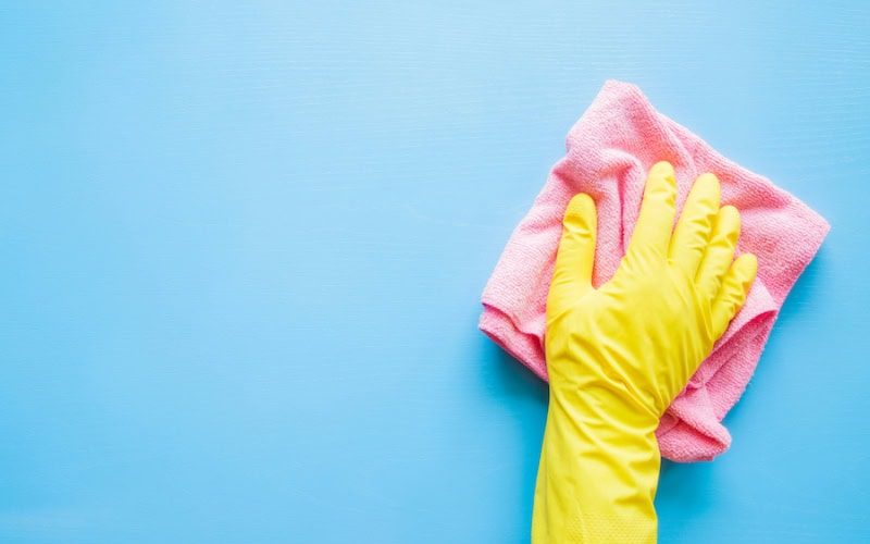 Photo of a hand in a yellow rubber glove, wiping a blue wall or table with a pink cloth.