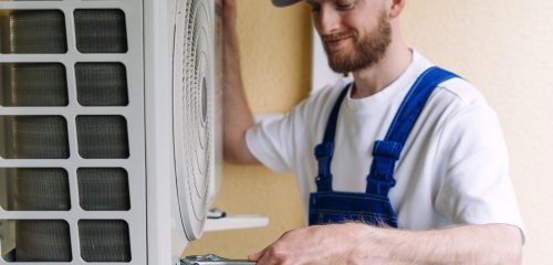An HVAC technician in blue overalls and a cap uses a wrench during an AC replacement in Picayune.
