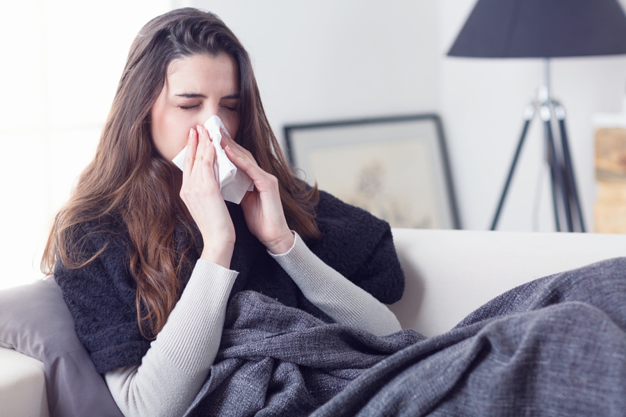 Young attractive woman lying sick at home couch in cold and flu sneezing nose with tissue covering with blanket. What Does Indoor Air Quality Have to Do With Allergies?